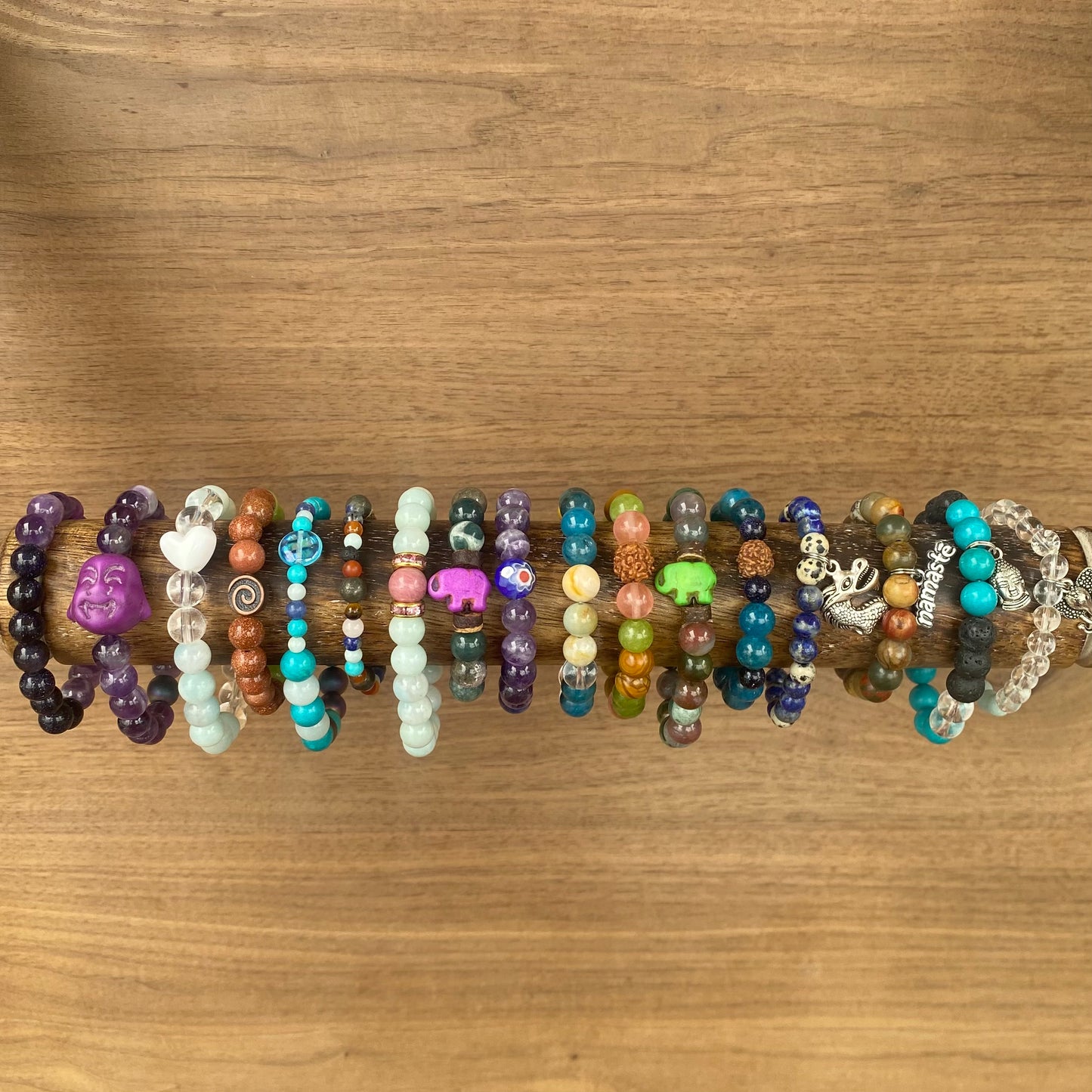 colorful beaded children's bracelets on a wooden rod, featuring a variety of gemstone beads, charms like a Buddha face, and small animal figures.